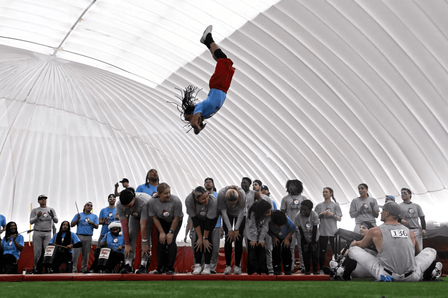 A tumbler somersaults high in the air above a line of teammates bent over side by side on the turf, inside an inflatable dome, while the rest of the squad and a drumline look on