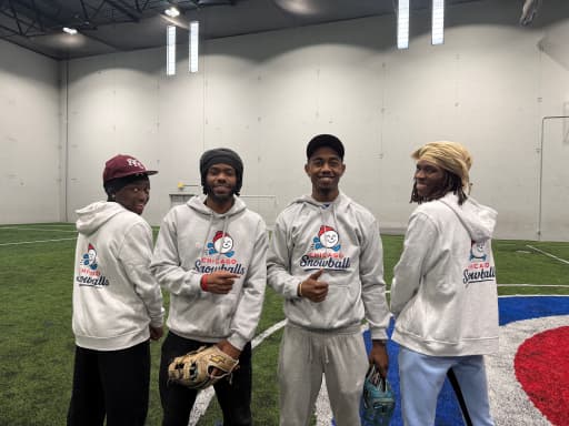 Four players in Rocket Squirrels hoodies stand shoulder to shoulder on the indoor turf, gloves in hand, smiling at the camera